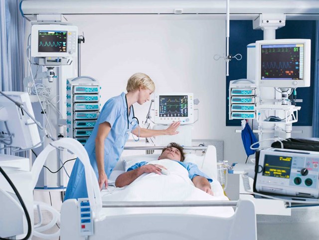 Female nurse checking on a sleeping male patient surrounded by medical devices and monitors.