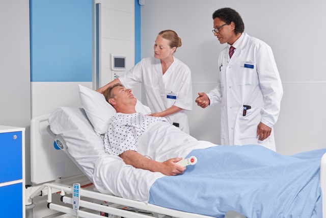 Male patient in a raised hospital bed, holding a pillow speaker in his right hand, attended by two medical professionals, a man and a womanMale patient in a raised hospital bed, holding a pillow speaker in his right hand, attended by two medical professionals, a man and a woman