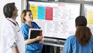 Two female nurses in blue scrubs and a male doctor in a white coat in front of a Digistat dashboard displayed on a big screen