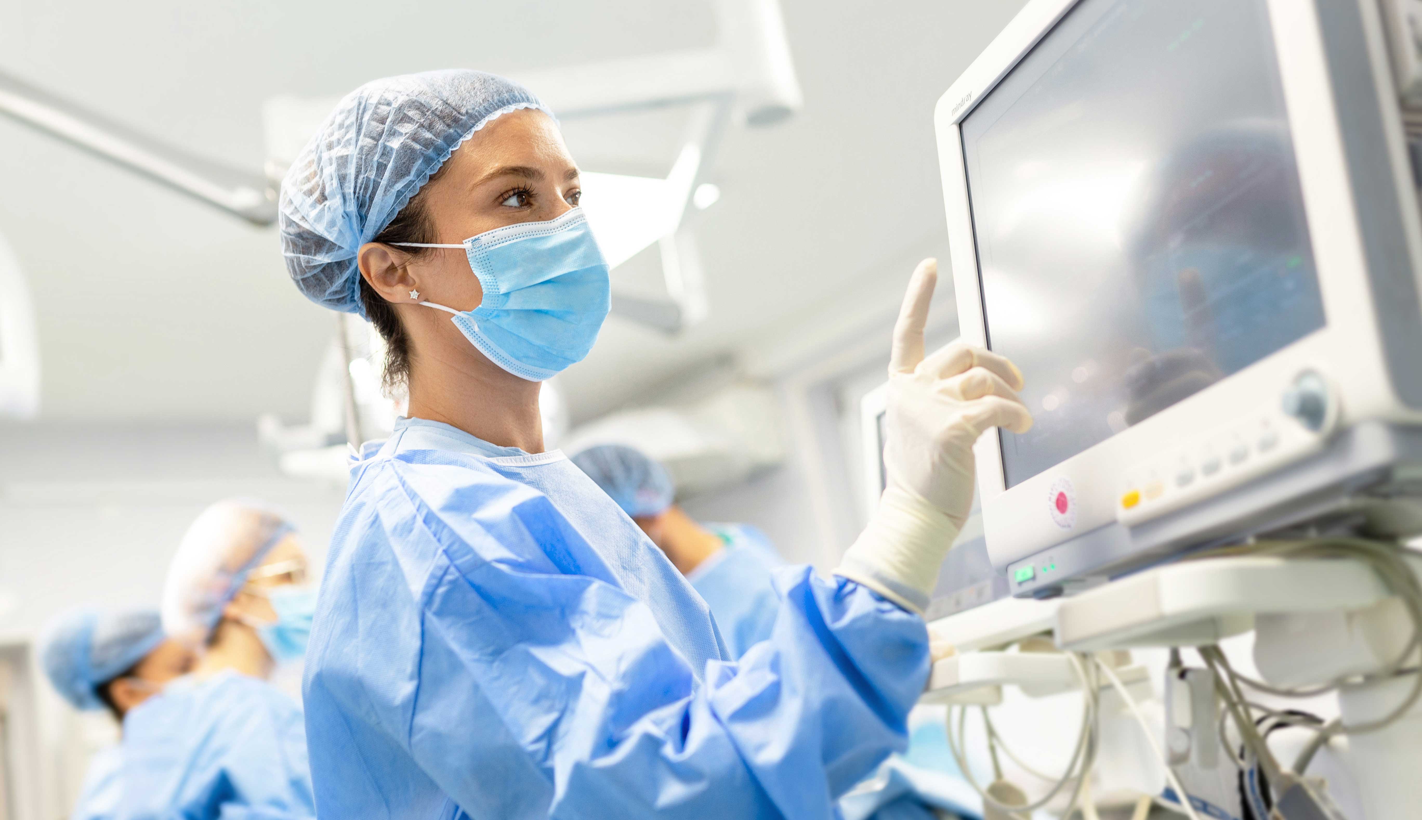 Close up of female medical personnel in PPE looking at a monitor, there are other members of the staff in the blurred background